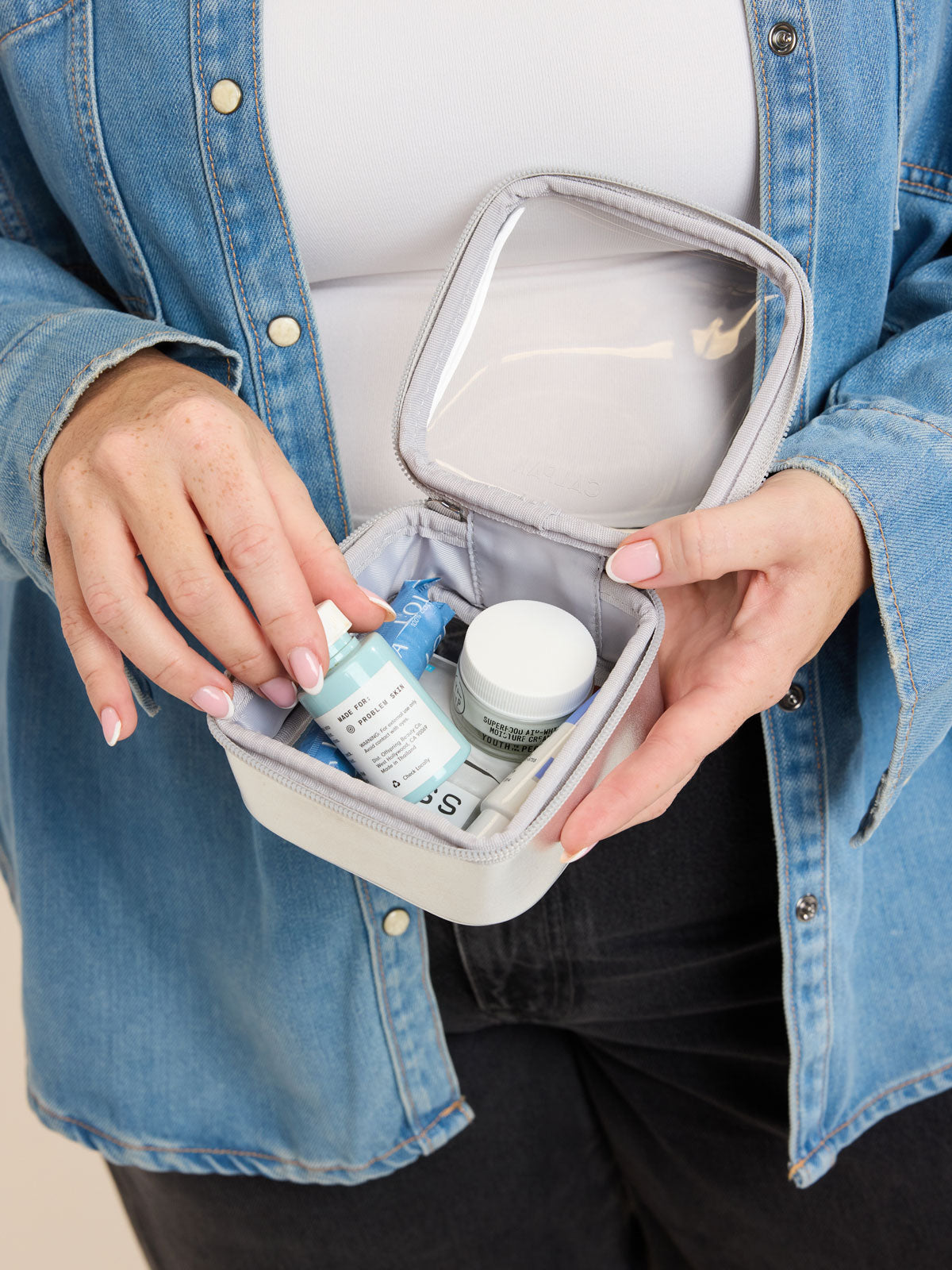 Model adding items into the CALPAK Tiny Clear Cosmetics Case in Silver; CCT2501-SILVER