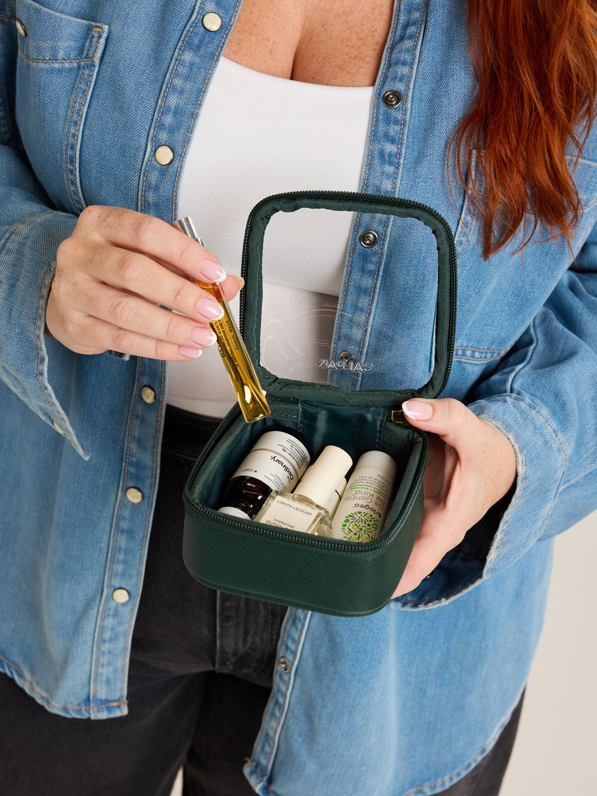 Model putting in items into CALPAK Tiny Clear Cosmetics Case in dark green Emerald on a white background; CCT2501-EMERALD
