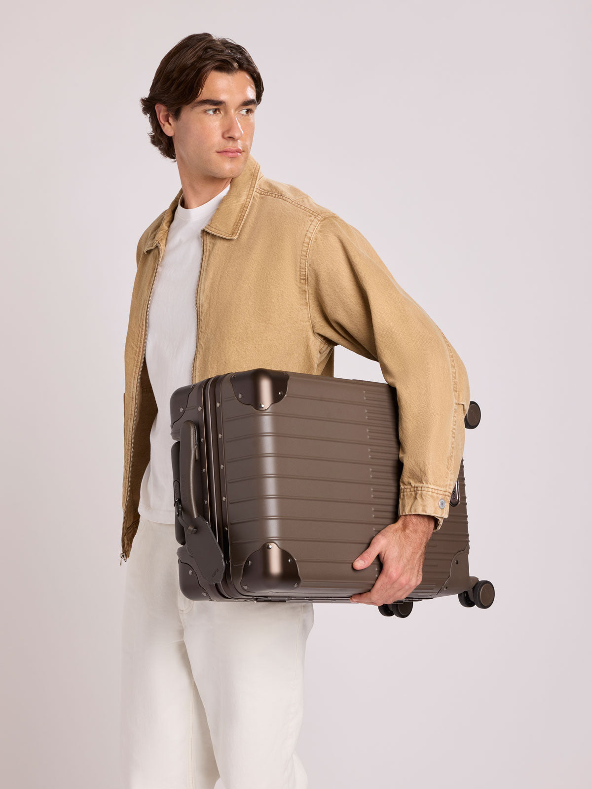 A man in a beige jacket and white shirt holds a CALPAK Vault brown Mushroom hard-shell spinner Carry On Luggage with a telescoping handle extended, featuring a black leatherette side handle and trim, set against a plain light gray background; LVT1020-MUSHROOM