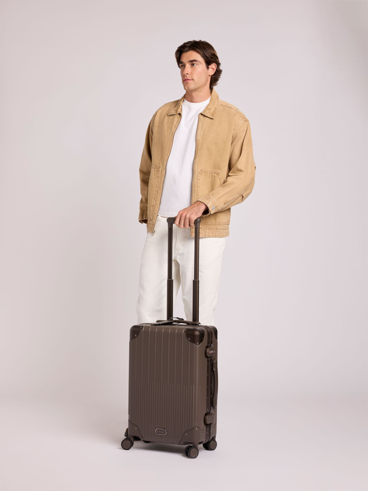 A man wearing a beige jacket, white t-shirt, and white pants stands next to a CALPAK Vault brown Mushroom hard-shell spinner Carry On Luggage with a telescoping handle extended, featuring a black leatherette side handle and trim, set against a plain light gray background; LVT1020-MUSHROOM