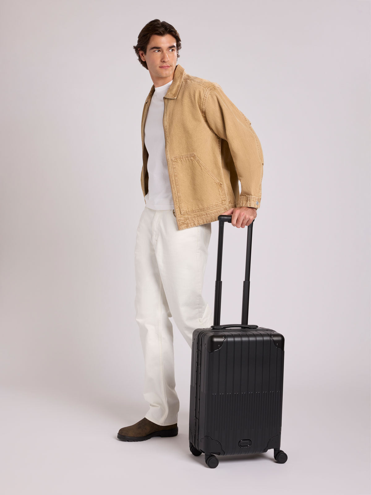 A man stands wearing a tan overshirt, white t-shirt, white trousers, and brown shoes, holding the handle of a CALPAK Vault black Onyx hard-shell spinner Carry On Luggage with a telescoping handle extended, featuring a Black leatherette side handle and trim, set against a plain light gray background;  LVT1020-ONYX