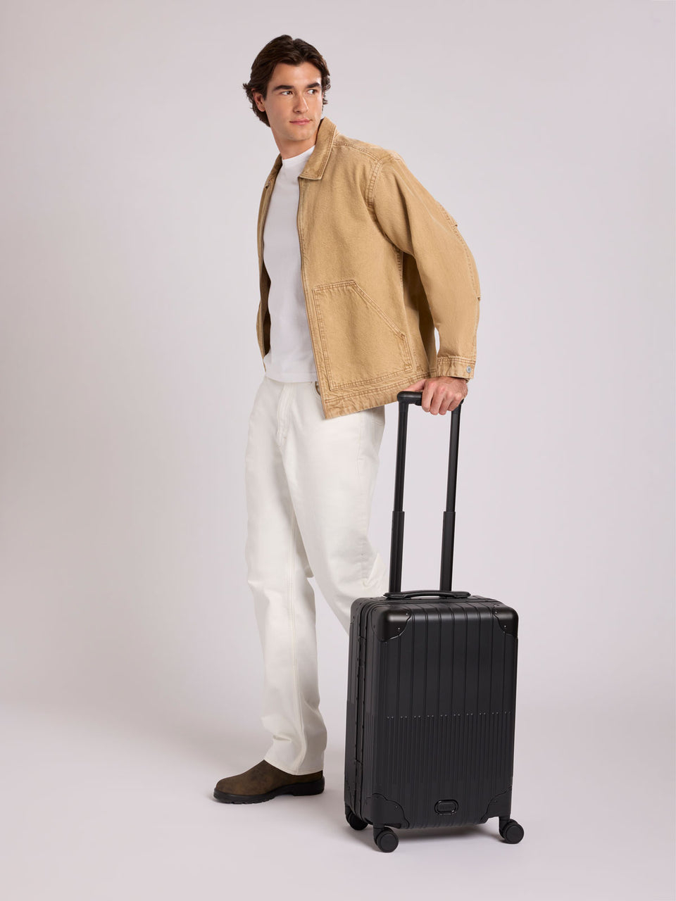 A man stands wearing a tan overshirt, white t-shirt, white trousers, and brown shoes, holding the handle of a CALPAK Vault black Onyx hard-shell spinner Carry On Luggage with a telescoping handle extended, featuring a Black leatherette side handle and trim, set against a plain light gray background;  LVT1020-ONYX