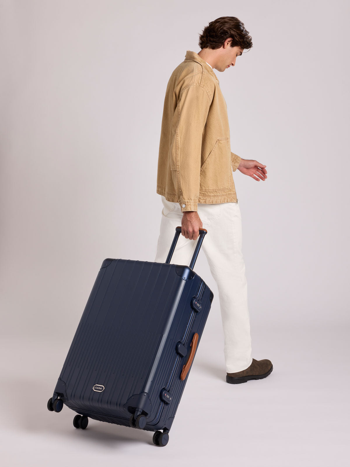 A man in a beige jacket and white pants walks while pulling a CALPAK Large Vault Luggage in blue Navy with hard-shell spinner suitcase with a ribbed vertical design, shown from the front on a light gray background; LVT1028-NAVY