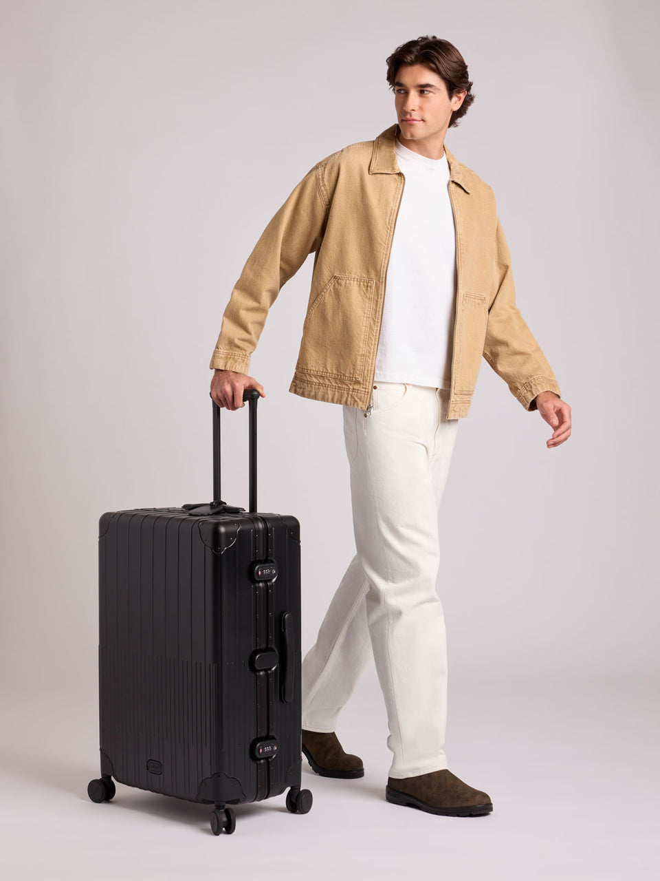 A man wearing a tan overshirt, white t-shirt, white trousers, and brown shoes stands next to a CALPAK Large Vault Luggage in black Onyx with hard-shell spinner suitcase with a ribbed vertical design, shown from the front on a light gray background; LVT1028-ONYX