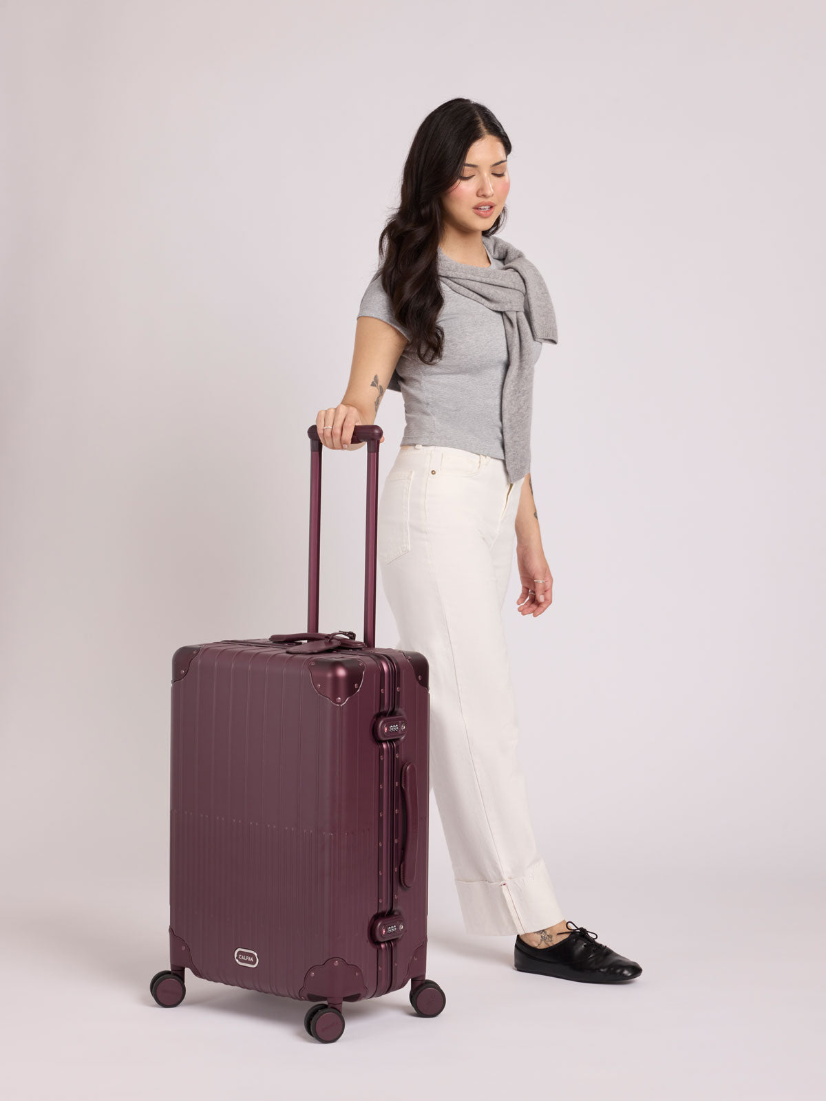 A woman wearing a grey top stands beside a CALPAK Vault Medium Luggage Dark Cherry deep red hard-shell spinner suitcase with a ribbed vertical design, shown from the front against a clean white background; LVT1024-DARK-CHERRY