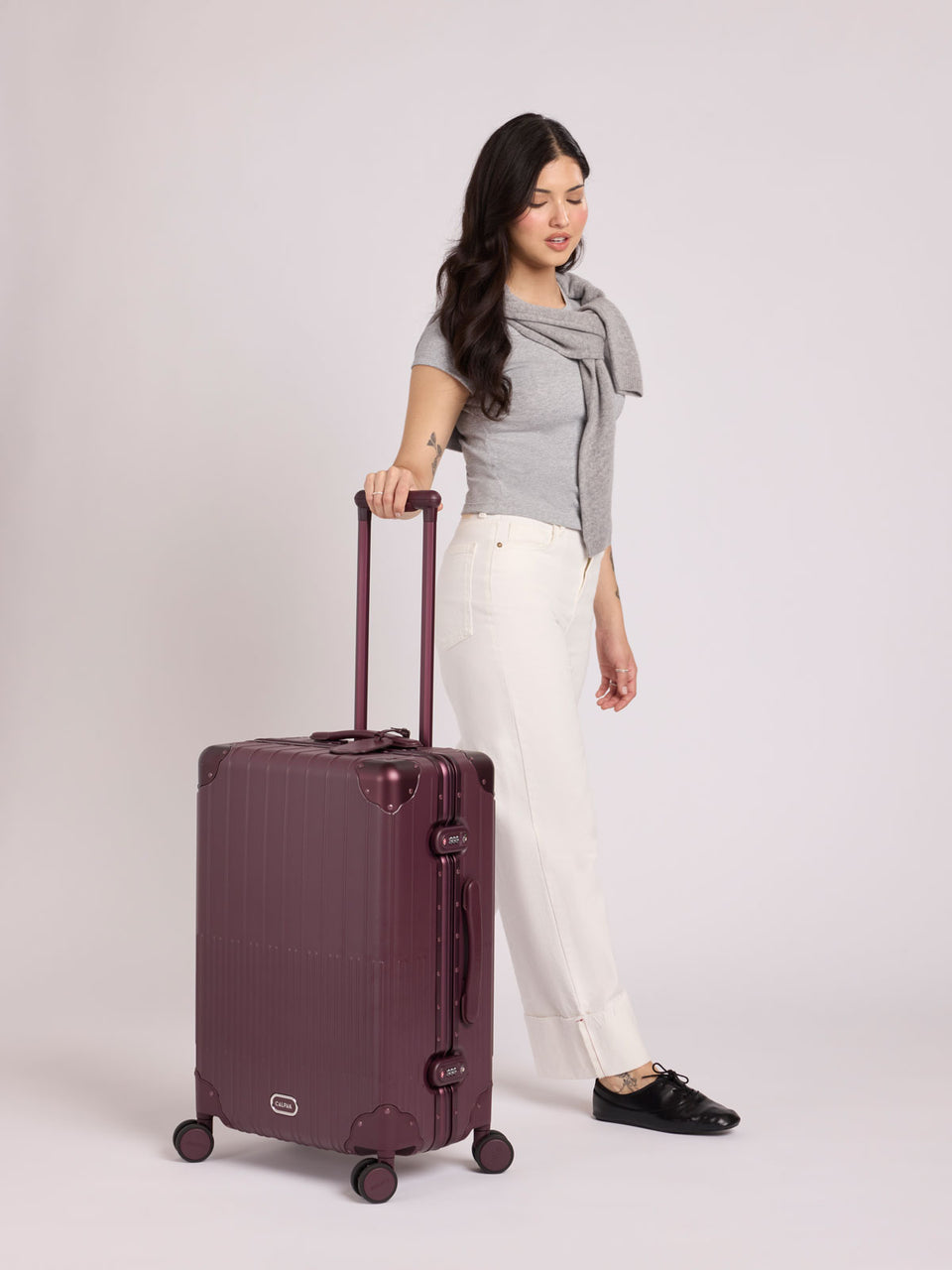 A woman wearing a grey top stands beside a CALPAK Vault Medium Luggage Dark Cherry deep red hard-shell spinner suitcase with a ribbed vertical design, shown from the front against a clean white background; LVT1024-DARK-CHERRY