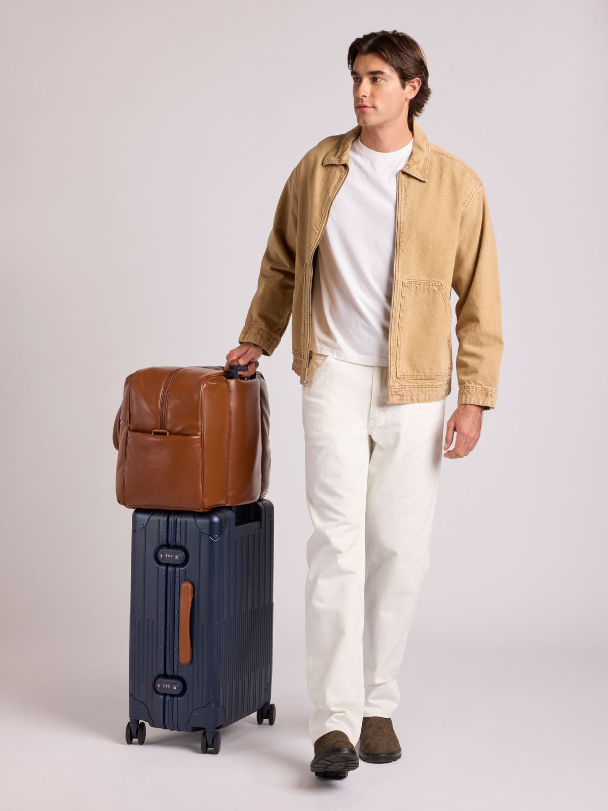 A man stands in a studio wearing a tan overshirt, white T-shirt, white trousers, and brown shoes, holding the handle of a CALPAK Vault Luggage blue Navy hard-shell spinner suitcase with a ribbed vertical design, shown from the front against a clean white background; LVT1024-NAVY