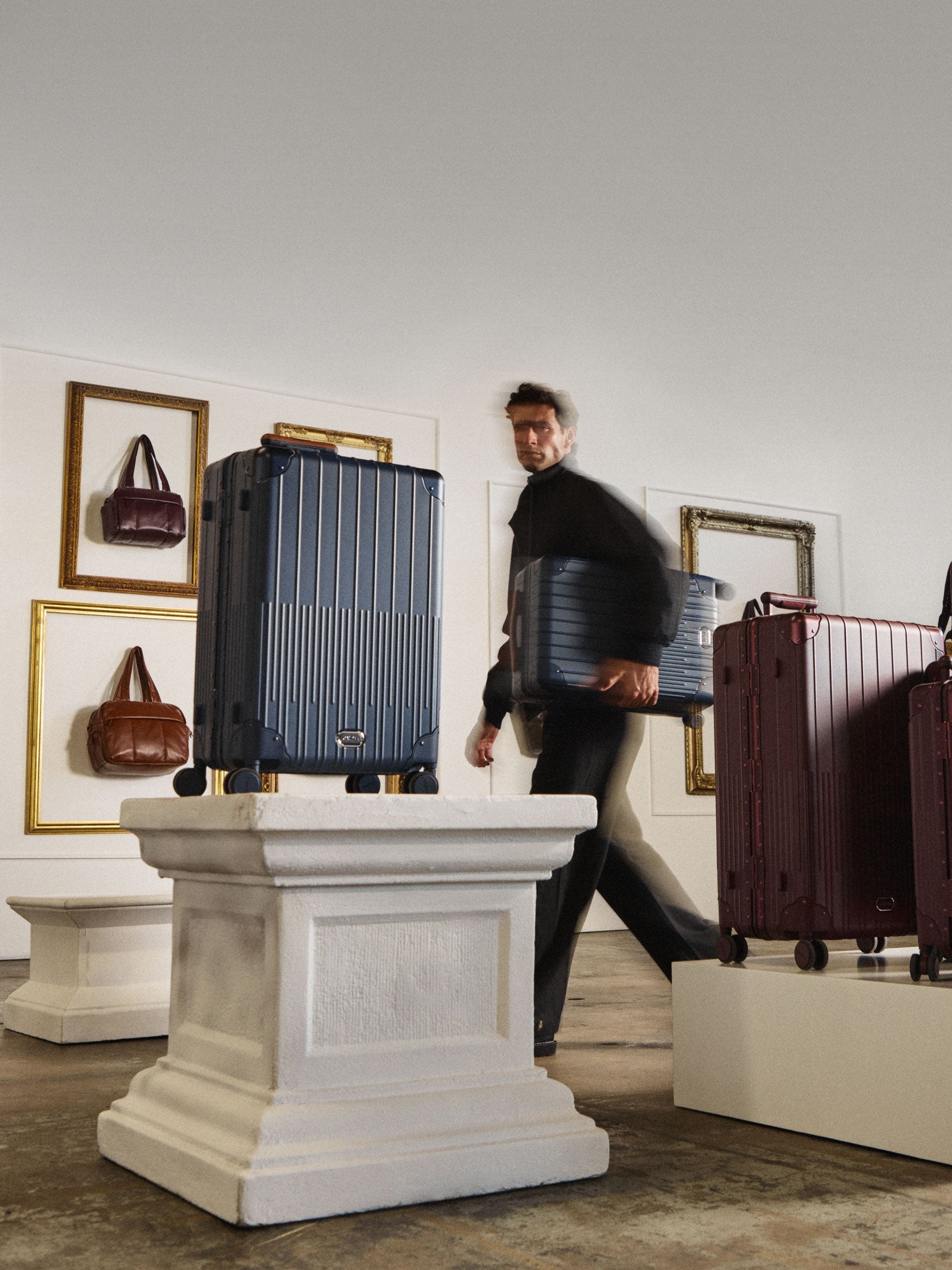 A man walks through a gallery-like interior carrying a blue hard-shell suitcase, with multiple hard-shell suitcases displayed on white pedestals in a room with framed wall art.