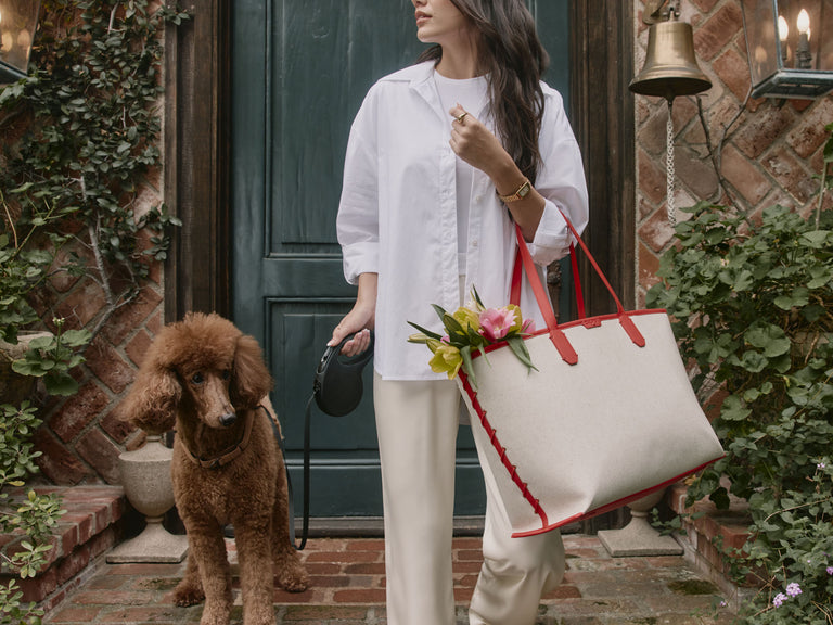 Woman holding a white CALPAK Colette Laptop tote bag with red handles and flowers, standing next to a brown dog outdoors.