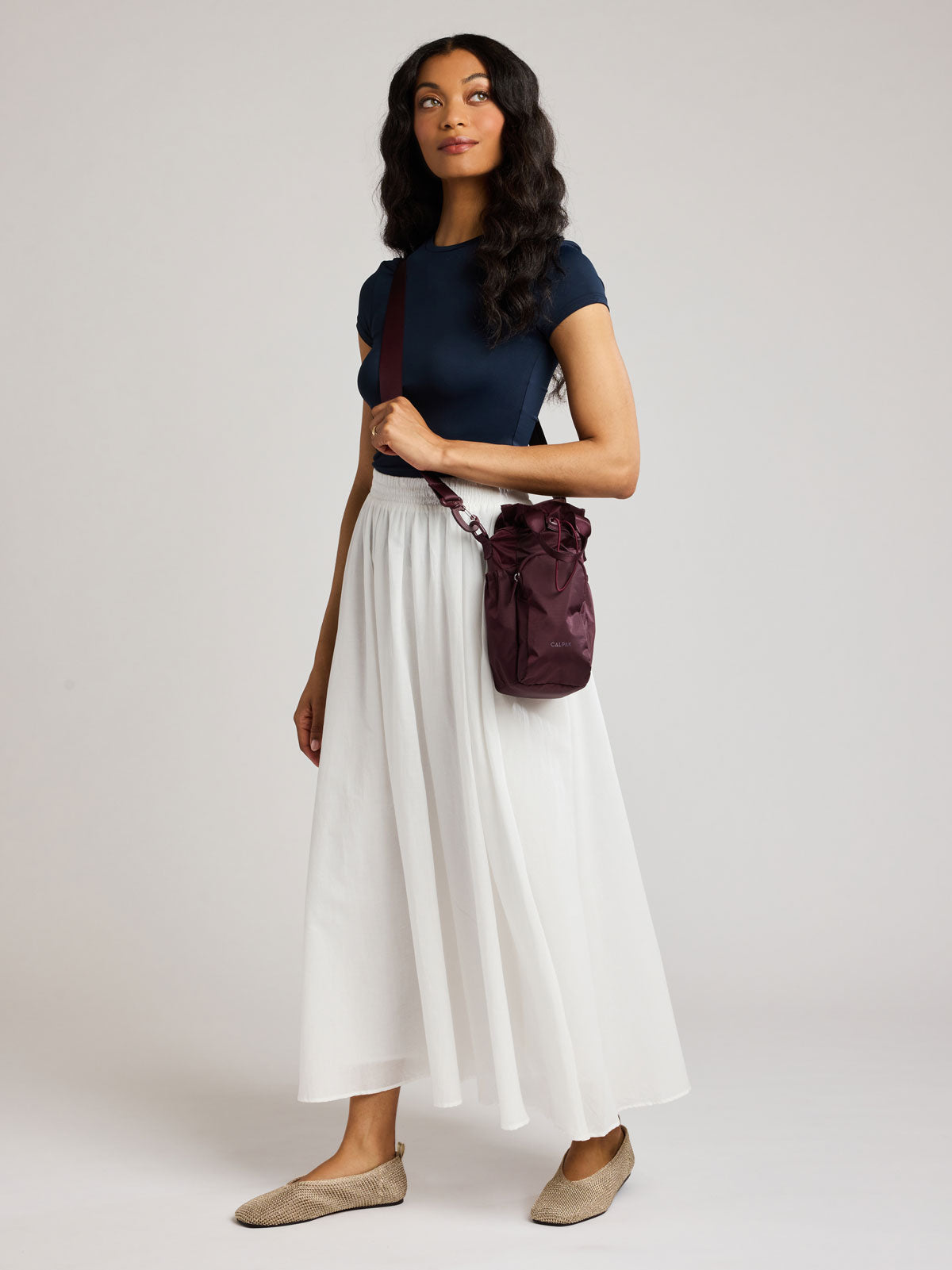 A woman modeling a navy blue short-sleeve top, a long white pleated skirt, and beige flats, carrying a dark burgundy CALPAK Water Bottle Holder with top handles, drawstring top, front zippered pocket, and a detachable shoulder strap; AWH2101-DARK-CHERRY