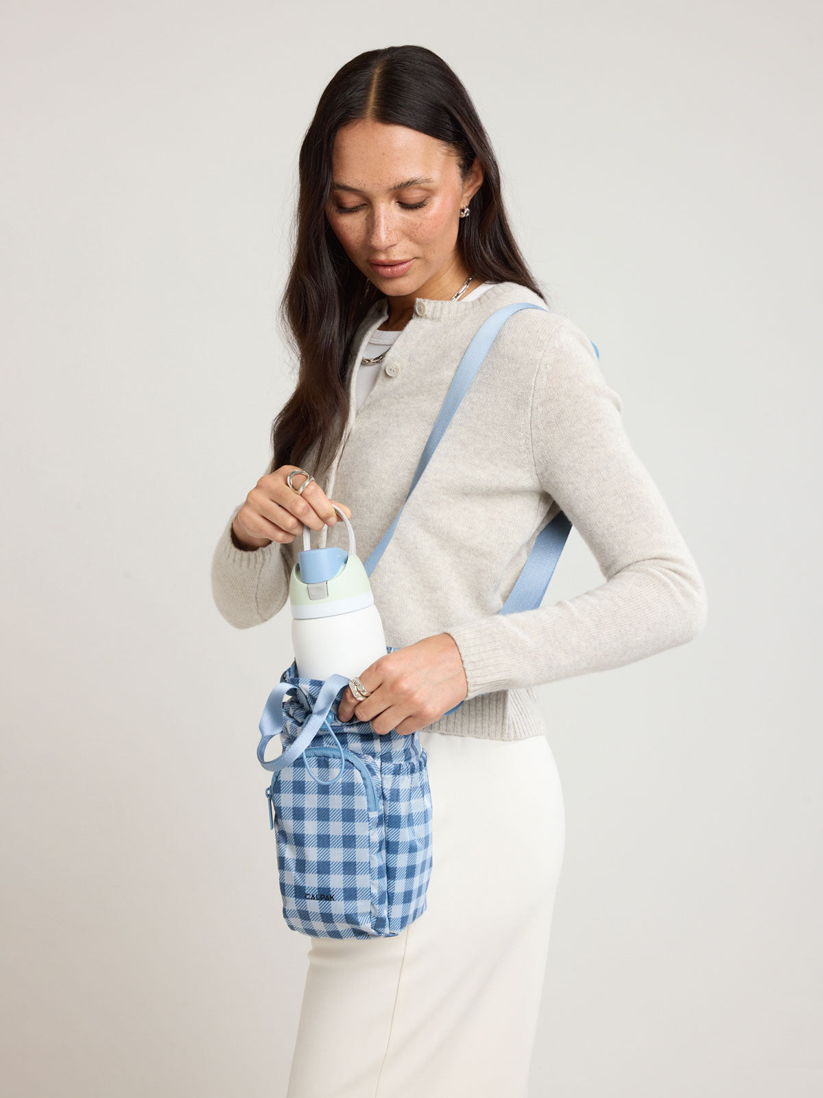 Woman putting a water bottle in the CALPAK Water Bottle Holder in blue and white checkered French Blue Gingham; AWH2101-FRENCH-BLUE-GINGHAM