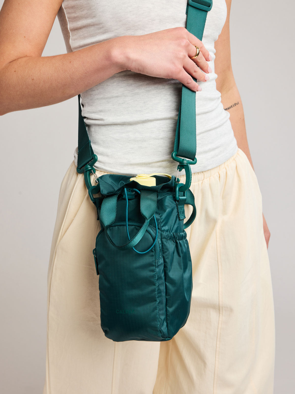 A person wearing a white tank top and cream pants carries a dark green teal Kale CALPAK Water Bottle Holder with top handles, drawstring top, front zippered pocket, and a detachable shoulder strap; AWH2101-KALE