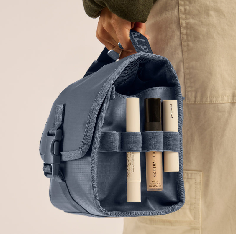 Person holding a blue CALPAK Terra Hanging Toiletry Bag with skincare products inside, wearing beige pants and a green cap